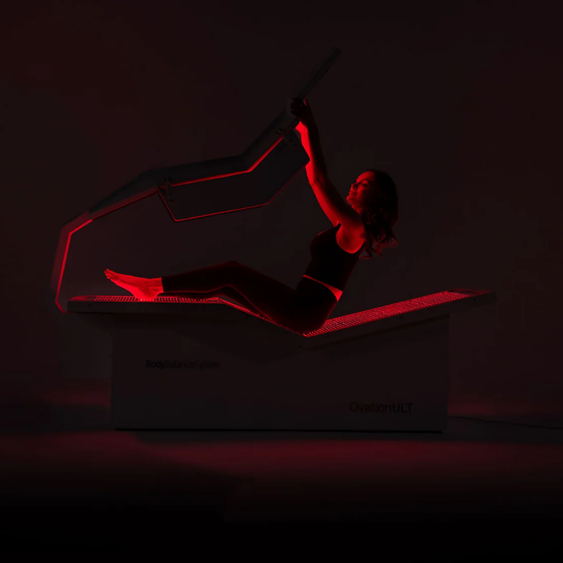Body Balance System Ovation ULT red light therapy bed on a black background with lights on and model.
